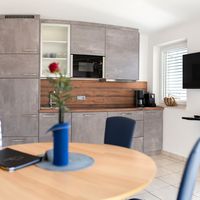 Helle Küche mit grauen Schränken, Holzrückwand, Esstisch und Fernseher am Fenster.
