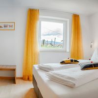 Helles Schlafzimmer mit großem Doppelbett, weißer Bettwäsche, gelben Vorhängen, Kleiderschrank und kleiner Holzbank.
