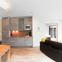 Offener Wohn- und Essbereich mit moderner Küche, Holztisch und schwarzem Sofa; durch die Glastür führt der Blick auf eine Terrasse mit Bergpanorama.