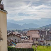 Blick über die Dächer eines Dorfes im Tal auf bewaldete Hügel und dahinterliegende Berge; links ist ein Balkon zu sehen.