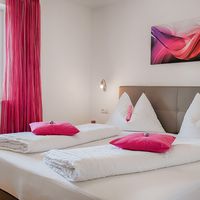 Ein ordentlich hergerichtetes Doppelbett mit weißer Bettwäsche und pinken Kissen steht neben einem Fenster mit pinken Vorhängen.