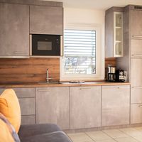 Moderne Küche mit grauen Einbauschränken, Holzarbeitsplatte, Spüle, Mikrowelle und Kaffeemaschinen; im Vordergrund steht ein Sofa.