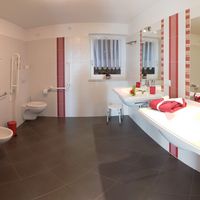 Helles Badezimmer mit ebenerdiger Dusche, WC, Bidet und langem Waschtisch mit großen Spiegeln; rote Handtücher und Fliesenstreifen setzen Farbakzente.