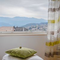 Blick aus einem offenen Fenster auf eine Stadt und dahinterliegende Berge; im Zimmer liegen ein grünes Kissen und gestreifte Vorhänge.
