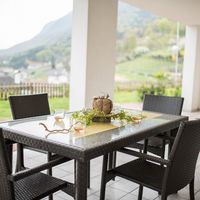 Ein gedeckter Gartentisch aus dunklem Flechtmaterial steht auf einer überdachten Terrasse mit Blick auf eine grüne Berglandschaft.