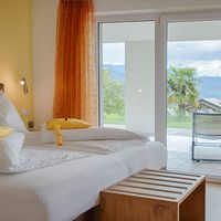 Helles Schlafzimmer mit weißem Bett, gelben Kissen und orangefarbenen Vorhängen; durch die Terrassentür sind Berge und ein Garten zu sehen.