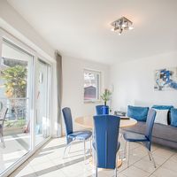 Heller Wohn- und Essbereich mit rundem Tisch, blauen Stühlen, grauem Sofa und großen Glastüren zum Balkon.