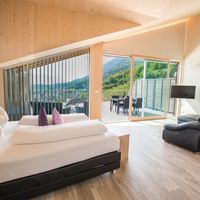Helles Schlafzimmer mit großem Bett, schwarzem Sofa und bodentiefen Fenstern mit Blick auf Berge und See.