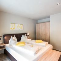 Helles Schlafzimmer mit Doppelbett, weißer Bettwäsche und gelben Kissen; daneben stehen ein Kleiderschrank und Nachttische.