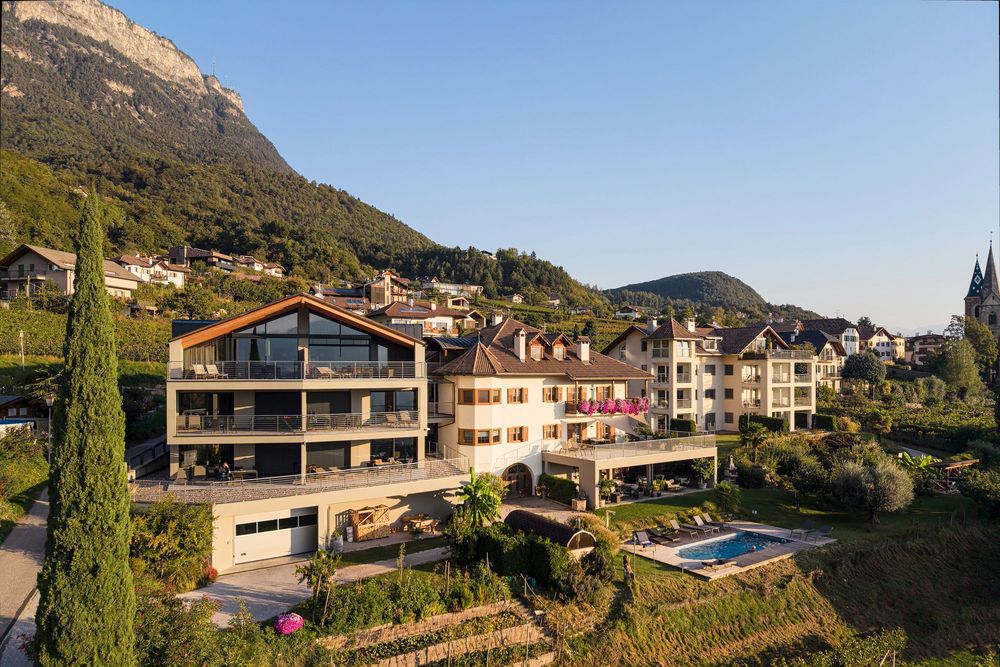 Südtiroler Dorf mit Häusern und Pool am Fuß eines bewaldeten Berges; rechts ist ein Kirchturm zu sehen.