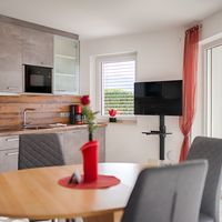 Heller Wohn- und Essbereich mit grauer Einbauküche, Esstisch, Fernseher und großer Glastür zur Terrasse.