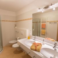 Helles Badezimmer mit Dusche, WC, Doppelwaschbecken und farbigen Handtüchern; durch die offene Tür ist ein Schlafzimmer zu sehen.