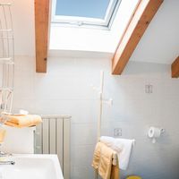 Helles Badezimmer unter dem Dach mit Dachfenster, Holzbalken, Waschbecken, Glaskabine und Toilette.