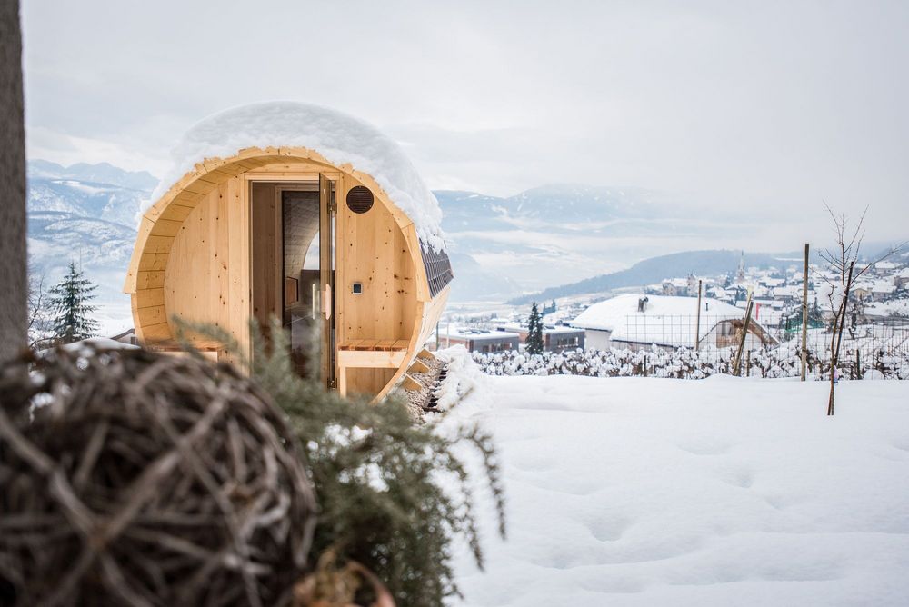Eine schneebedeckte, tonnenförmige Holzhütte steht vor einer winterlichen Berglandschaft mit einem Dorf im Tal.