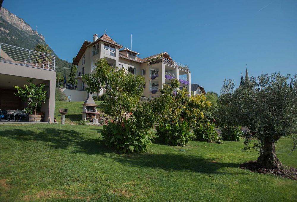 Mehrstöckiges Wohnhaus mit Balkonen und blühenden Pflanzen in einem gepflegten Garten vor einem bewaldeten Berg.