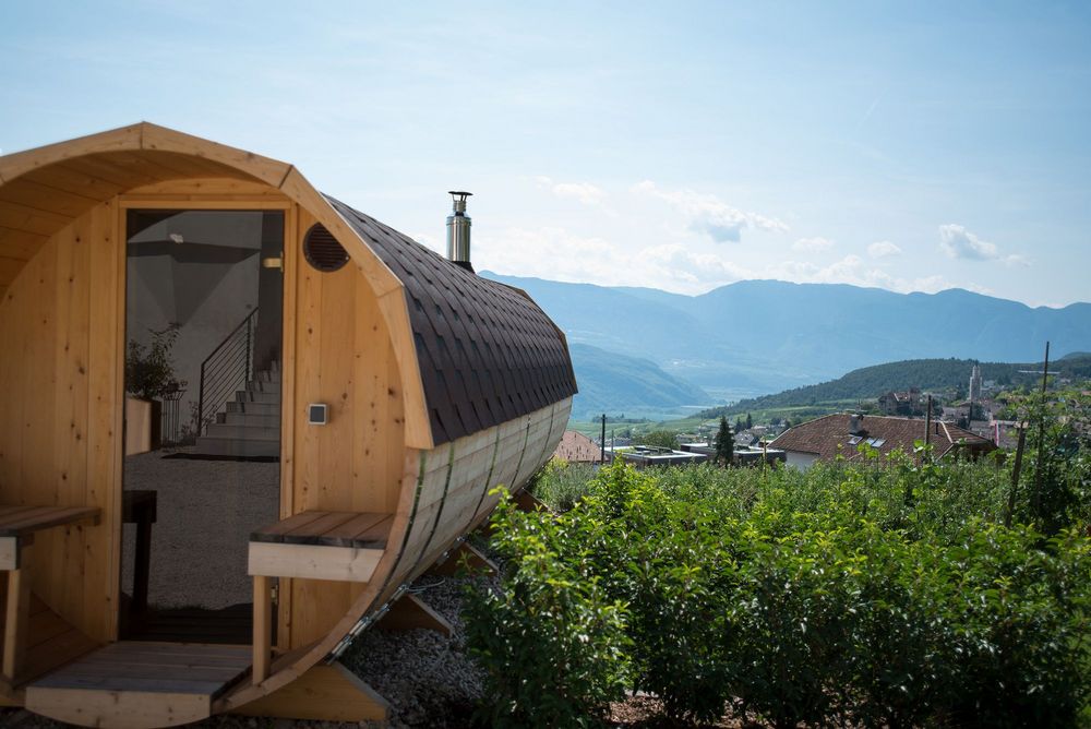 Eine moderne, tonnenförmige Holzhütte steht vor einer grünen Berglandschaft mit einem Dorf im Tal.