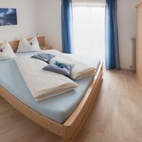 Helles Schlafzimmer mit einem großen Holzbett, weißer Bettwäsche, blauem Vorhang und Kleiderschrank aus Holz.