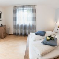 Helles Schlafzimmer mit Doppelbett, weißen Kissen, grauen Vorhängen und einer Holzkommode.