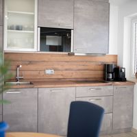 Moderne Küche mit grauen Schränken, Holzarbeitsplatte, Spüle, Kochfeld und Kaffeemaschinen neben einem Fenster.