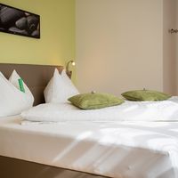 Ein ordentlich gemachtes Doppelbett mit weißer Bettwäsche und grünen Kissen steht in einem hellen Schlafzimmer.