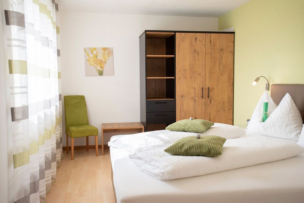 Helles Schlafzimmer mit weiß bezogenem Doppelbett, grünen Kissen, Holzschrank und grünem Stuhl am Fenster.