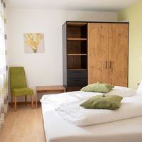Helles Schlafzimmer mit weiß bezogenem Doppelbett, grünen Kissen, Holzschrank und grünem Stuhl am Fenster.