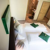 Modernes Hotelzimmer mit zwei nebeneinanderstehenden Betten, weißen Decken und grünen Kissen; im Hintergrund liegt ein holzverkleidetes Bad.