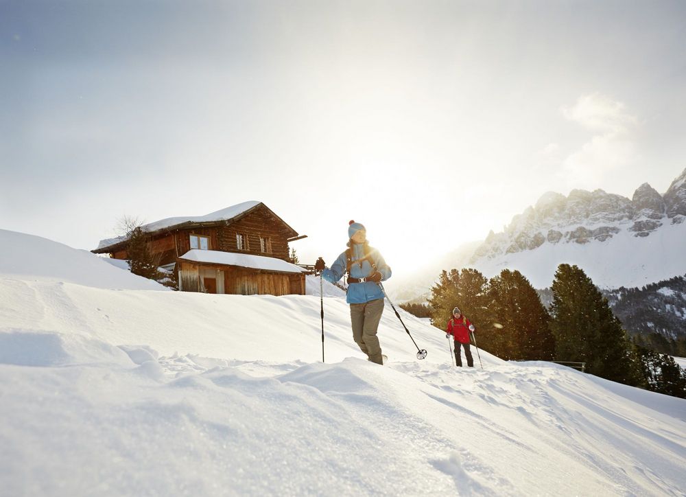 Zwei Personen wandern mit Skistöcken durch verschneite Berge; neben ihnen steht eine Holzhütte, dahinter leuchtet die tief stehende Sonne.