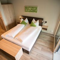 Modernes Schlafzimmer mit Doppelbett, grünen Kissen, Holzfußboden und offenem Blick ins Bad sowie zur Balkontür.