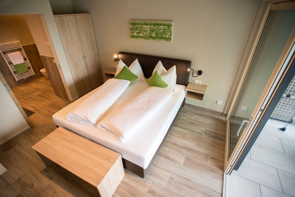 Modernes Schlafzimmer mit Doppelbett, grünen Kissen, Holzfußboden und offenem Blick ins Bad sowie zur Balkontür.