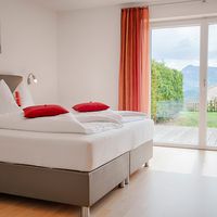 Helles Schlafzimmer mit weiß bezogenem Doppelbett und roten Kissen; durch die großen Glastüren blickt man auf Garten und Berge.