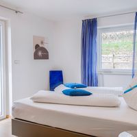 Helles Schlafzimmer mit weiß bezogenem Doppelbett, blauen Kissen und Vorhängen sowie einer Glastür zur Terrasse.