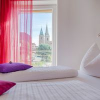 Helles Schlafzimmer mit zwei weiß bezogenen Betten und pinken Kissen; durch rote Vorhänge sind zwei Kirchtürme zu sehen.