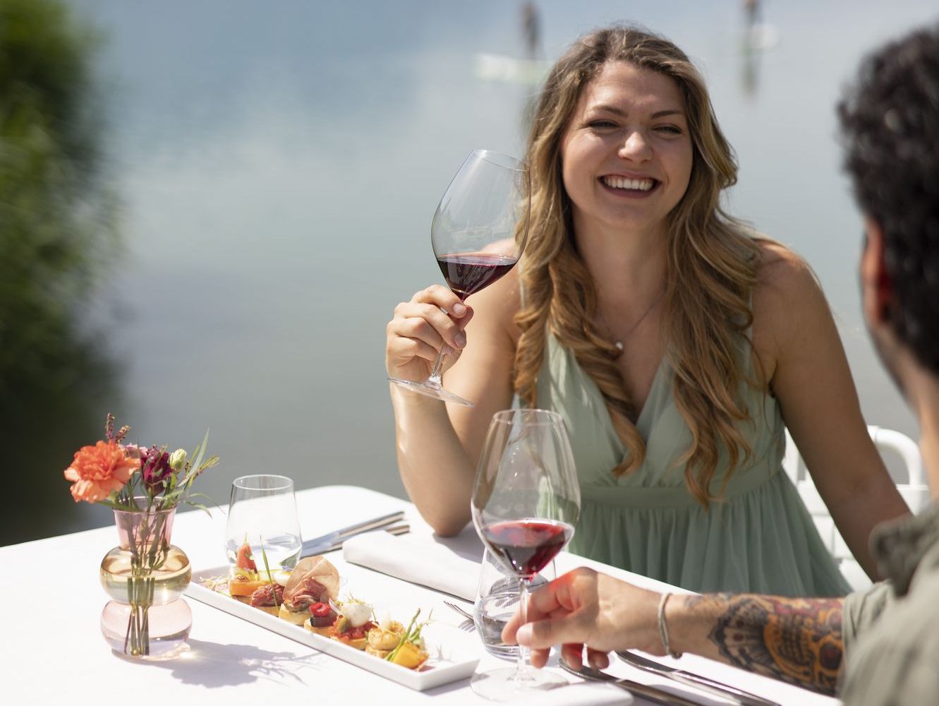 Eine lächelnde Frau hebt bei einem Essen am See ein Glas Rotwein; auf dem Tisch stehen kleine Speisen und Blumen.