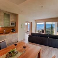 Offener Wohn- und Küchenbereich mit schwarzem Sofa, Holztisch und großen Glastüren zu einer Terrasse mit Bergblick.