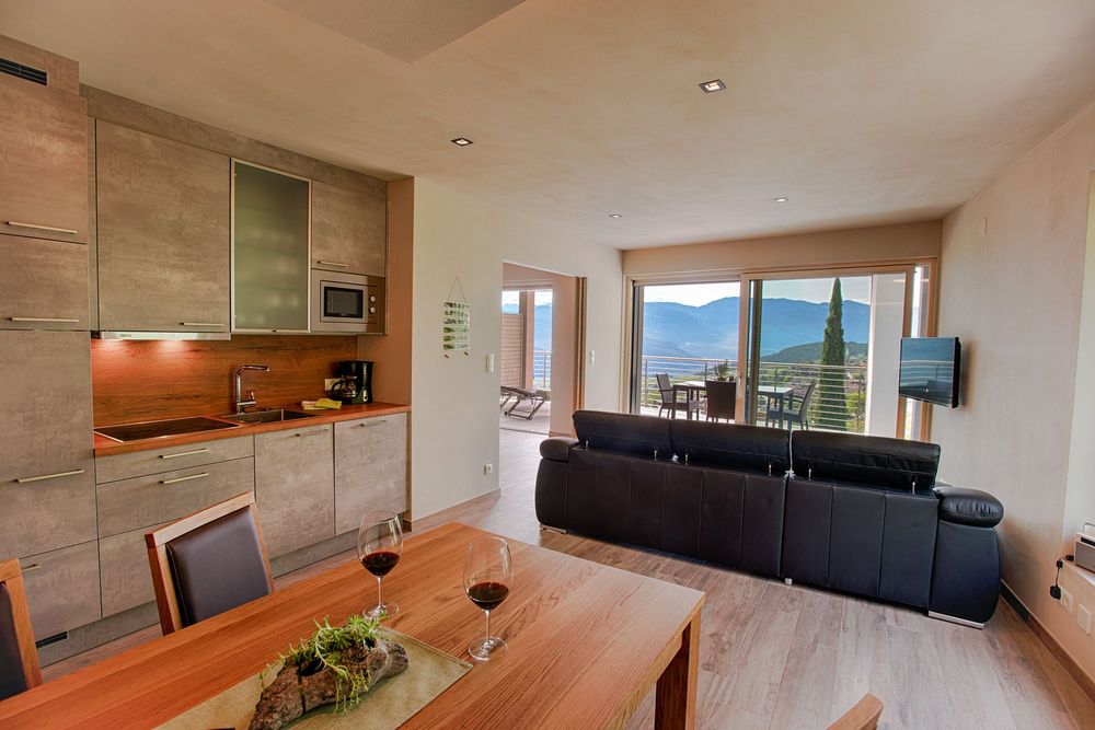Offener Wohn- und Küchenbereich mit schwarzem Sofa, Holztisch und großen Glastüren zu einer Terrasse mit Bergblick.