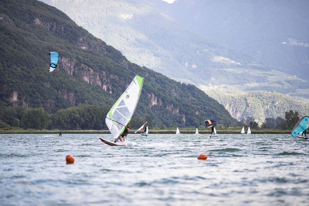 Mehrere Menschen surfen mit Segeln und Foilboards auf einem See vor bewaldeten Bergen.