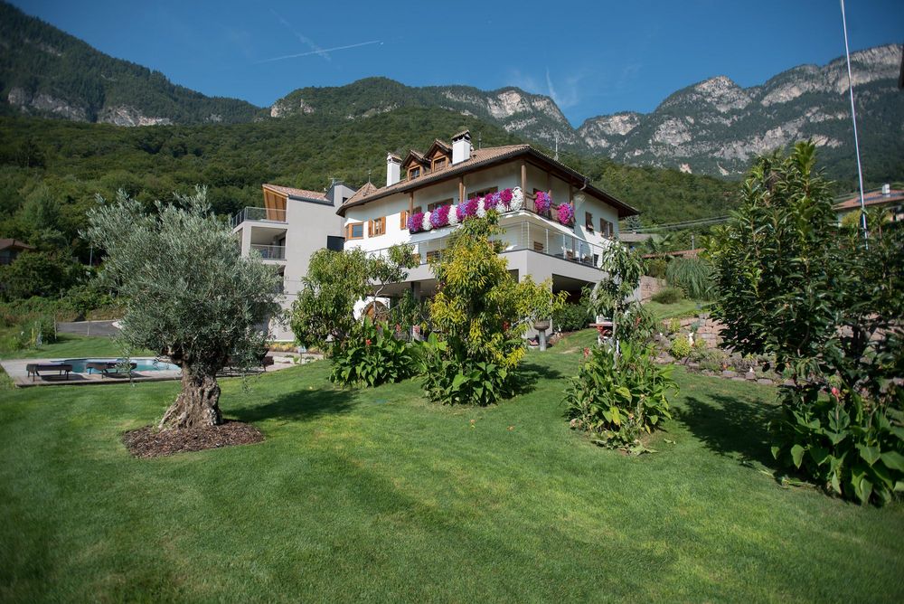 Ein Haus mit blumengeschmücktem Balkon steht in einem gepflegten Garten mit Pool vor bewaldeten Bergen.