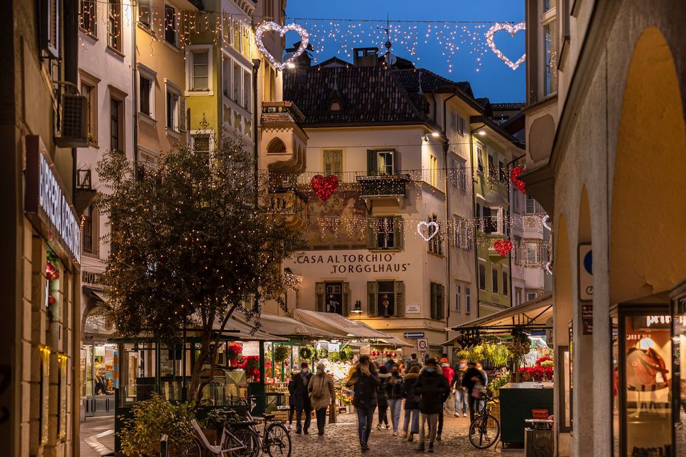 Belebte Altstadtgasse bei Dämmerung mit Marktständen, warmen Lichterketten und leuchtenden Herzen über den Häusern.