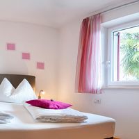 Helles Schlafzimmer mit Doppelbett, weißen Kissen, pinken Zierkissen und Blick auf eine Palme am Fenster.