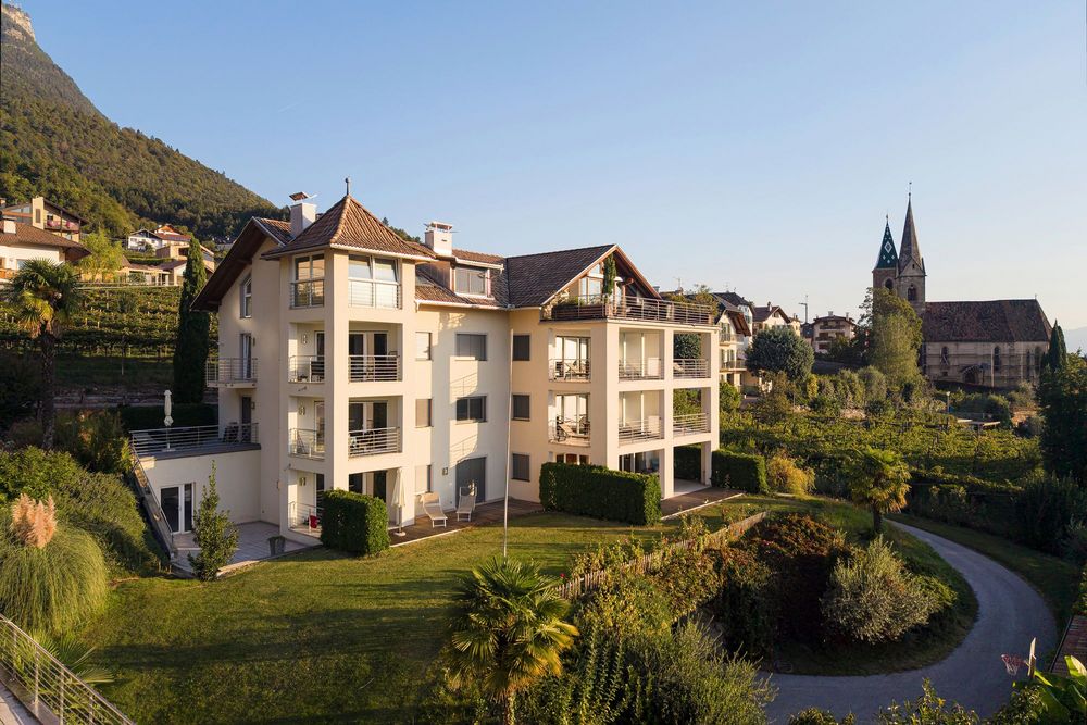 Modernes Wohnhaus mit Balkonen und Garten vor einem bewaldeten Berghang; rechts steht eine Kirche mit zwei spitzen Türmen.