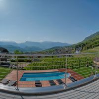Panoramablick von einer sonnigen Terrasse mit Pool auf ein Dorf und bewaldete Berge.