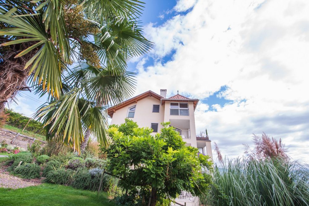 Ein helles Wohnhaus mit rotem Ziegeldach steht in einem üppig begrünten Garten unter einem wolkigen Himmel; links ragen Palmwedel ins Bild.