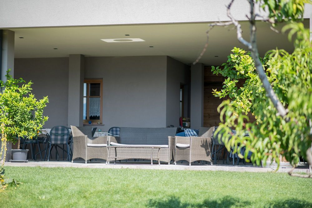 Überdachte Terrasse mit Korbsesseln, Sofa und Gartentischen, umgeben von grünen Sträuchern und Rasen.