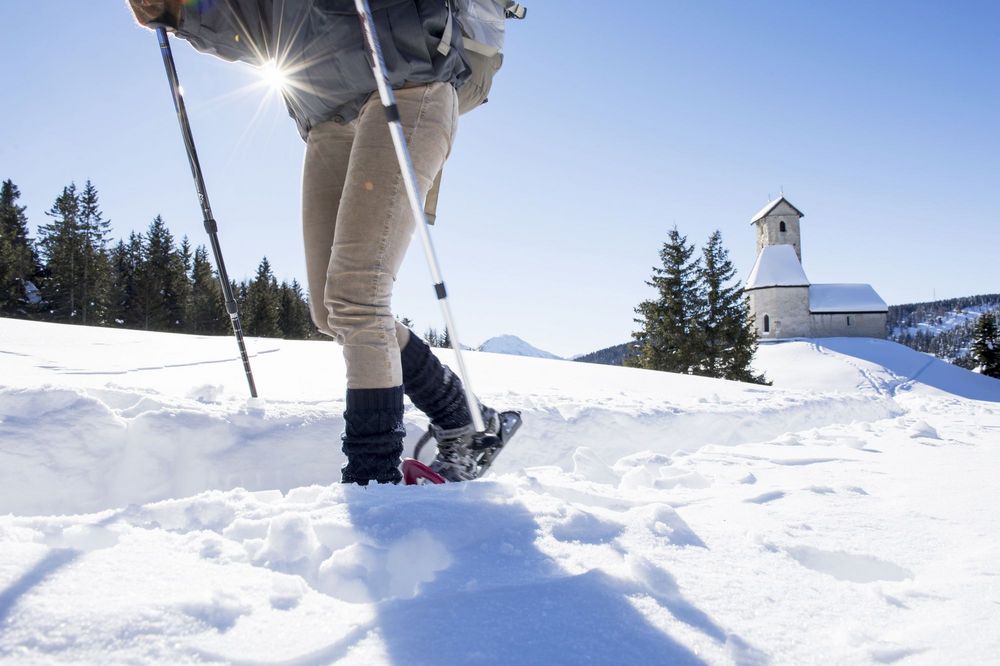 Eine Person wandert mit Schneeschuhen und Stöcken durch eine verschneite Landschaft; im Hintergrund steht eine kleine Kirche.