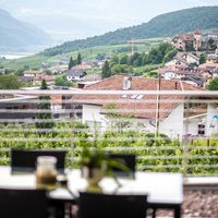 Blick durch ein Fenster auf ein Dorf mit Kirchturm, Weinbergen und bewaldeten Bergen; im Vordergrund sind unscharf ein Tisch und Stühle zu sehen.