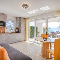 Heller Wohn- und Essbereich mit grauer Küchenzeile, orangefarbenen Stühlen und Sofa sowie großen Glastüren zu einer Terrasse mit Bergblick.