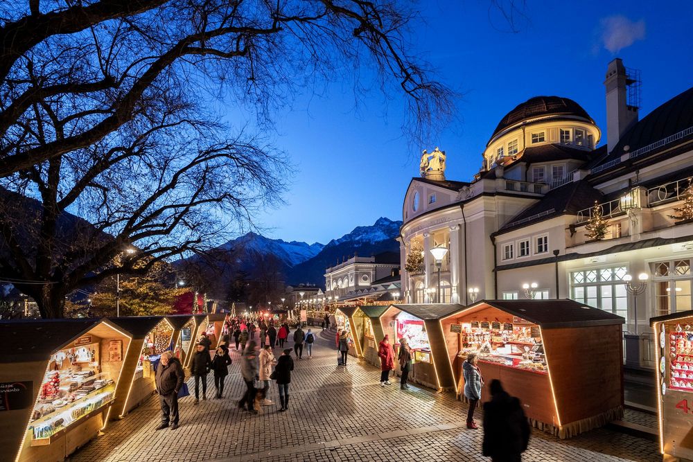 Belebter Weihnachtsmarkt mit beleuchteten Holzständen vor einem historischen Gebäude und schneebedeckten Bergen in der blauen Abenddämmerung.
