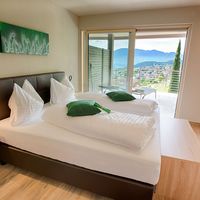 Helles Schlafzimmer mit großem Bett und grünen Kissen; durch die Balkontür sind ein Ort und Berge zu sehen.