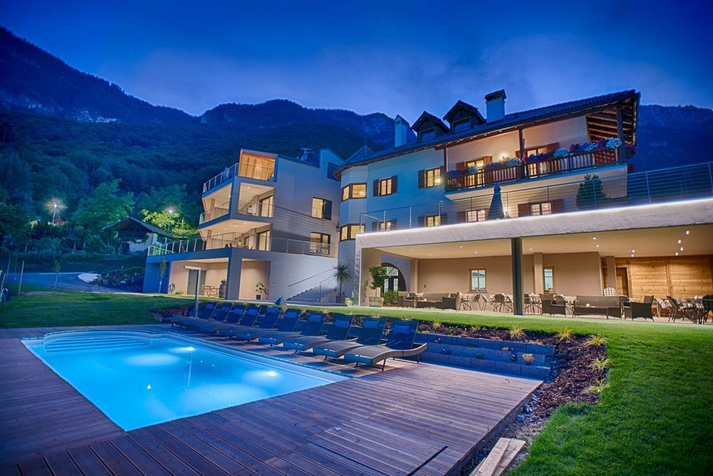 Ein beleuchtetes Hotel mit Balkon und Terrasse steht nachts vor bewaldeten Bergen; davor liegen ein Pool und mehrere Liegestühle.
